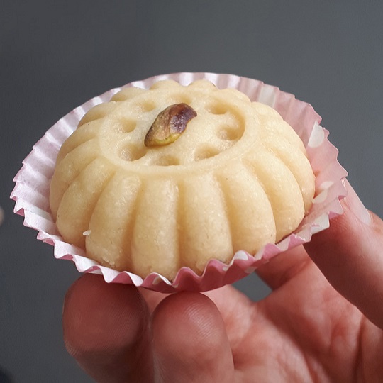 Photo close up of a biscuit in Tasmans hand, biscuit is pale off white colour and is shaped in a circle like shape with indents from a mould, it is like an old fashioned jelly mould shape , the biscuit has a brown and green pistachio placed ontop